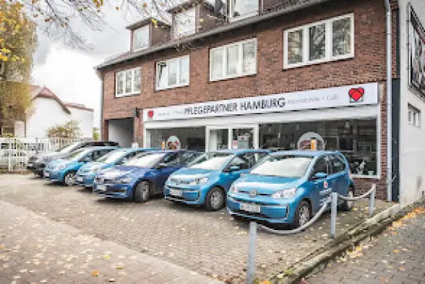 Eine Reihe von fünf blauen Autos steht geparkt vor einem zweistöckigen Backsteingebäude mit der Aufschrift "Pflegepartner Hamburg". Im Hintergrund sind Bäume und ein bewölkter Himmel zu sehen.