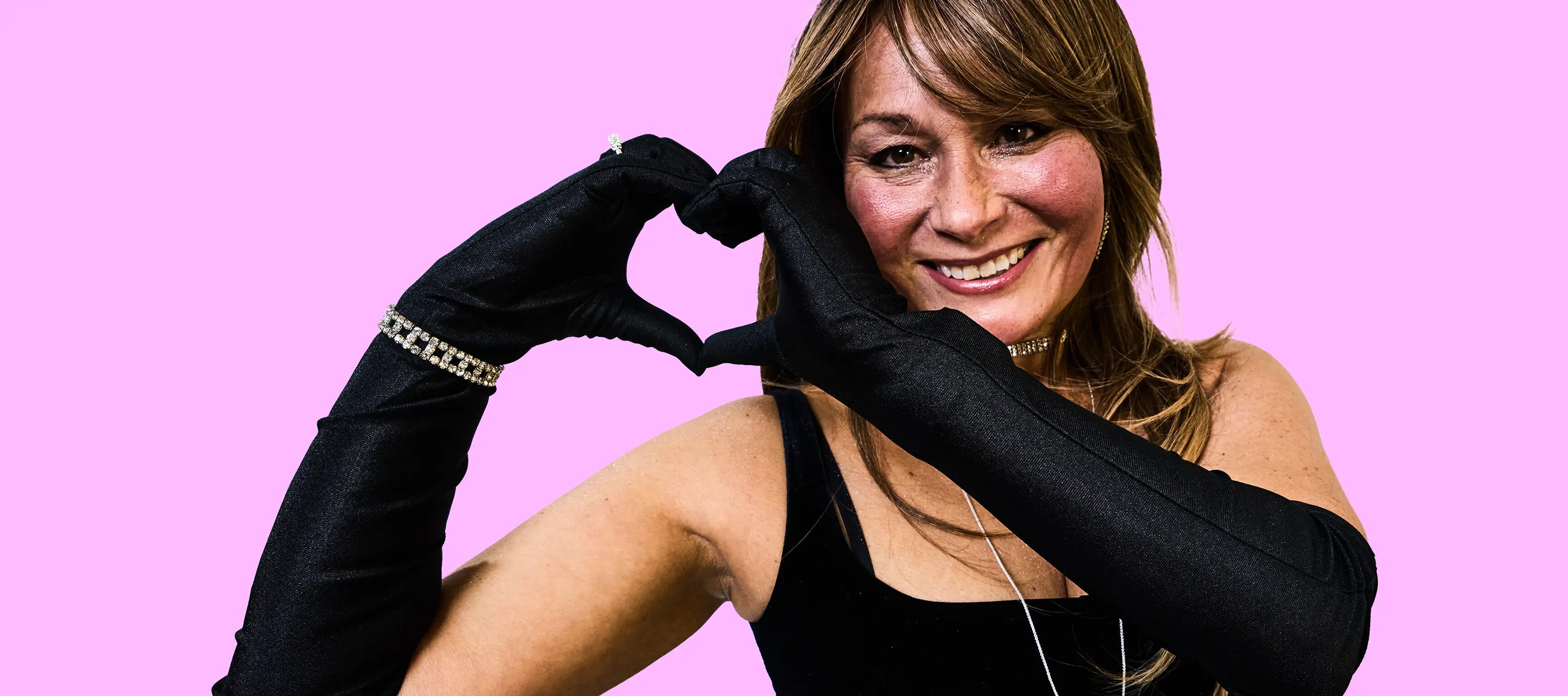 Eine lächelnde Person mit langen Haaren formt mit beiden Händen ein Herz. Sie trägt schwarze lange Handschuhe mit Schmuck und ein schwarzes Oberteil. Der Hintergrund ist pink.