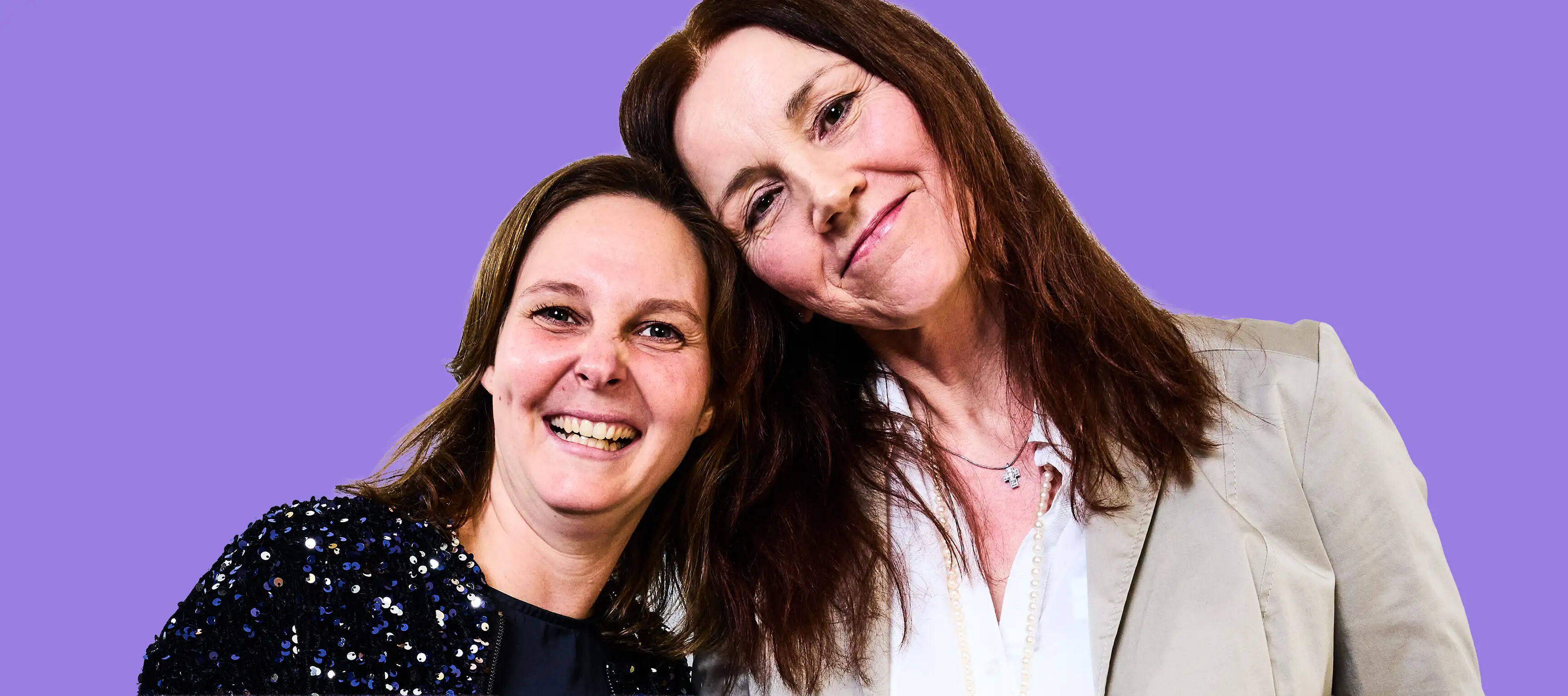 Zwei Personen stehen nah beieinander und lächeln. Die linke Person trägt eine Jacke mit Pailletten, die rechte Person eine helle Bluse und einen Blazer. Der Hintergrund ist einfarbig lila.
