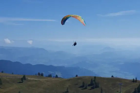 Eine Person fliegt an einem bunten Gleitschirm über eine hügelige Landschaft mit Bäumen. Der Himmel ist blau und klar, und im Hintergrund sind Bergketten zu sehen.