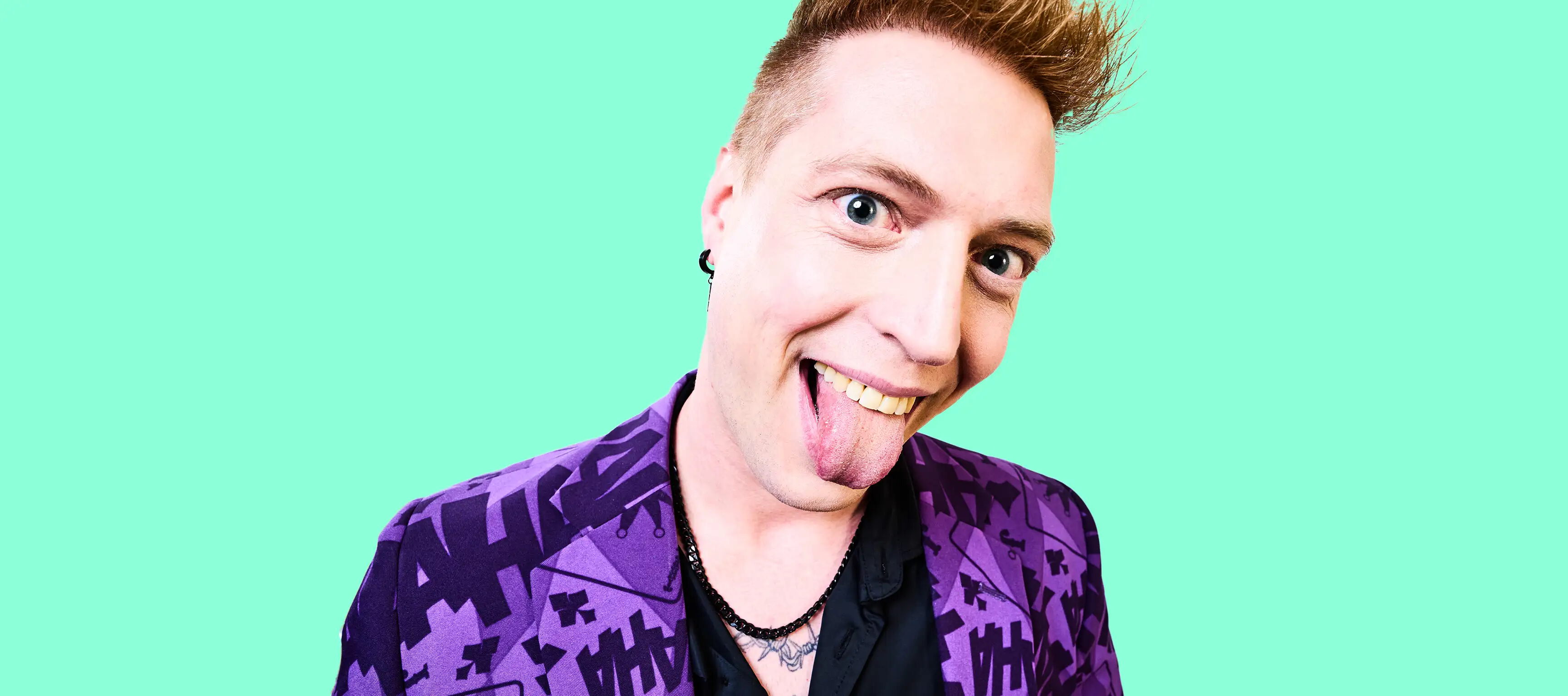 Ein lächelnder Mensch mit hochgestylten Haaren trägt einen lilafarbenen Anzug mit schwarzen Mustern. Der Hintergrund ist türkis. Die Person zeigt frech die Zunge.