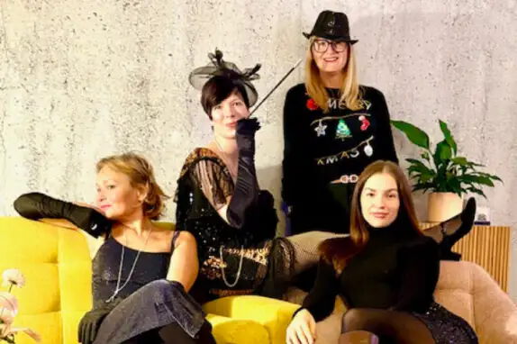 Vier Personen posieren auf einem gelben Sofa vor einer grauen Wand. Drei tragen elegante Abendkleidung und lange Handschuhe, eine Person trägt einen Weihnachts-Pullover und einen Hut. Neben dem Sofa steht eine Pflanze.