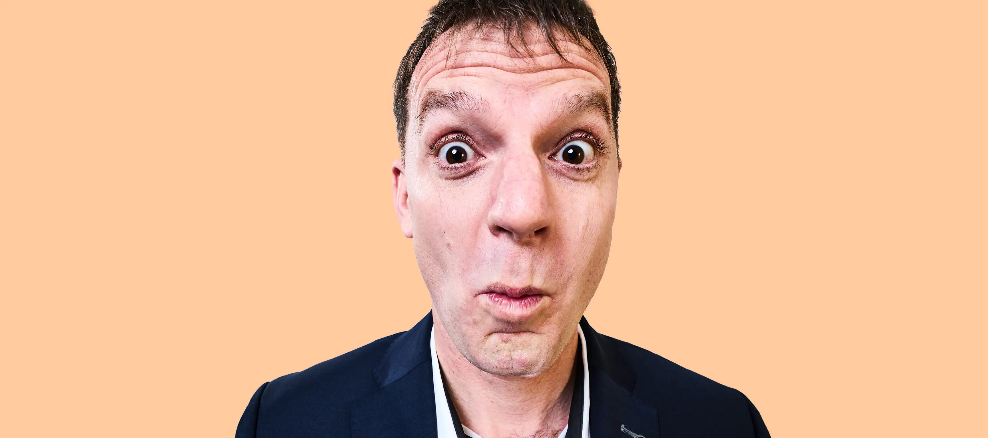 Eine Person in einem dunklen Sakko und mit kurzen Haaren schaut direkt in die Kamera. Der Hintergrund ist einfarbig orange. Die Person hat einen überraschten Gesichtsausdruck.