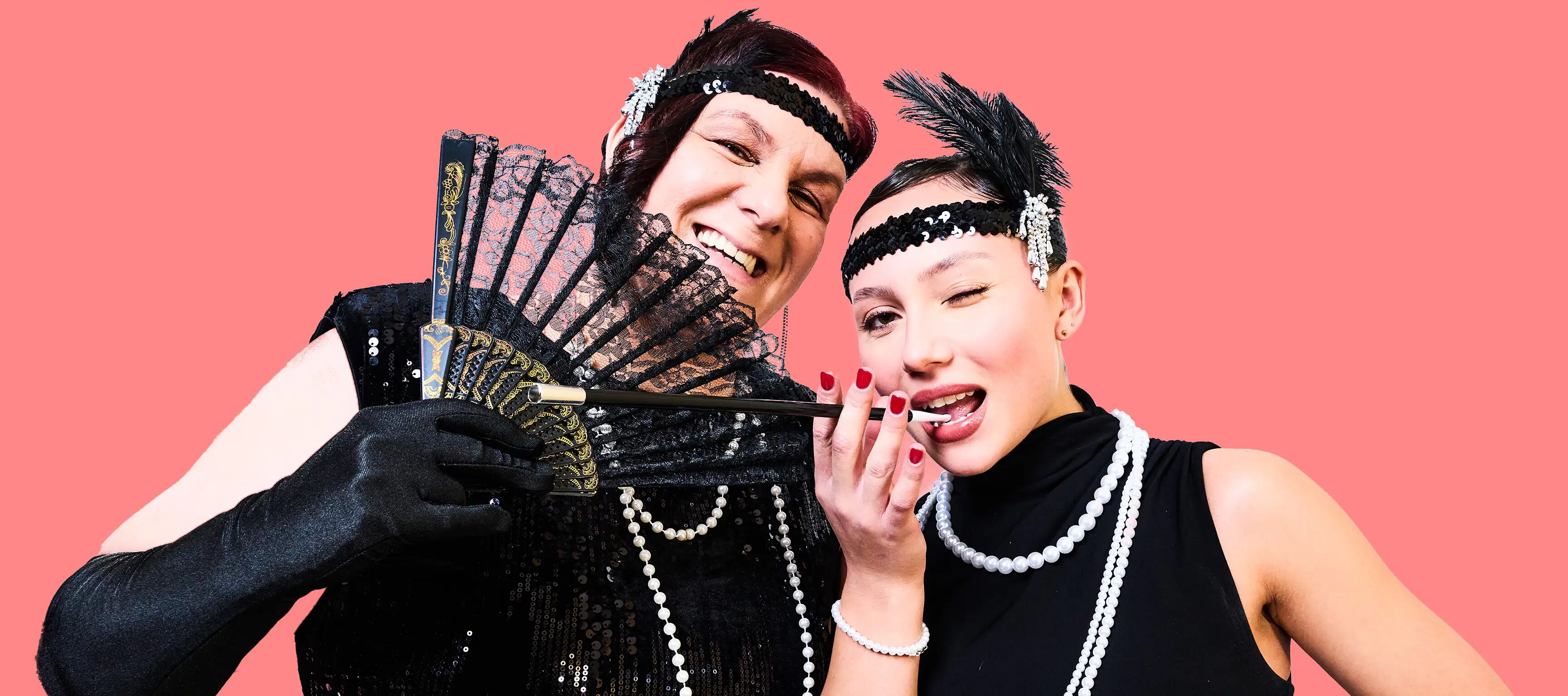 Zwei Personen in Retro-Kleidung posieren lächelnd vor einem pinken Hintergrund. Beide tragen Stirnbänder mit Federn, Perlenketten und schwarze Kleidung. Eine Person hält einen Fächer, die andere ein Zigarettenmundstück.