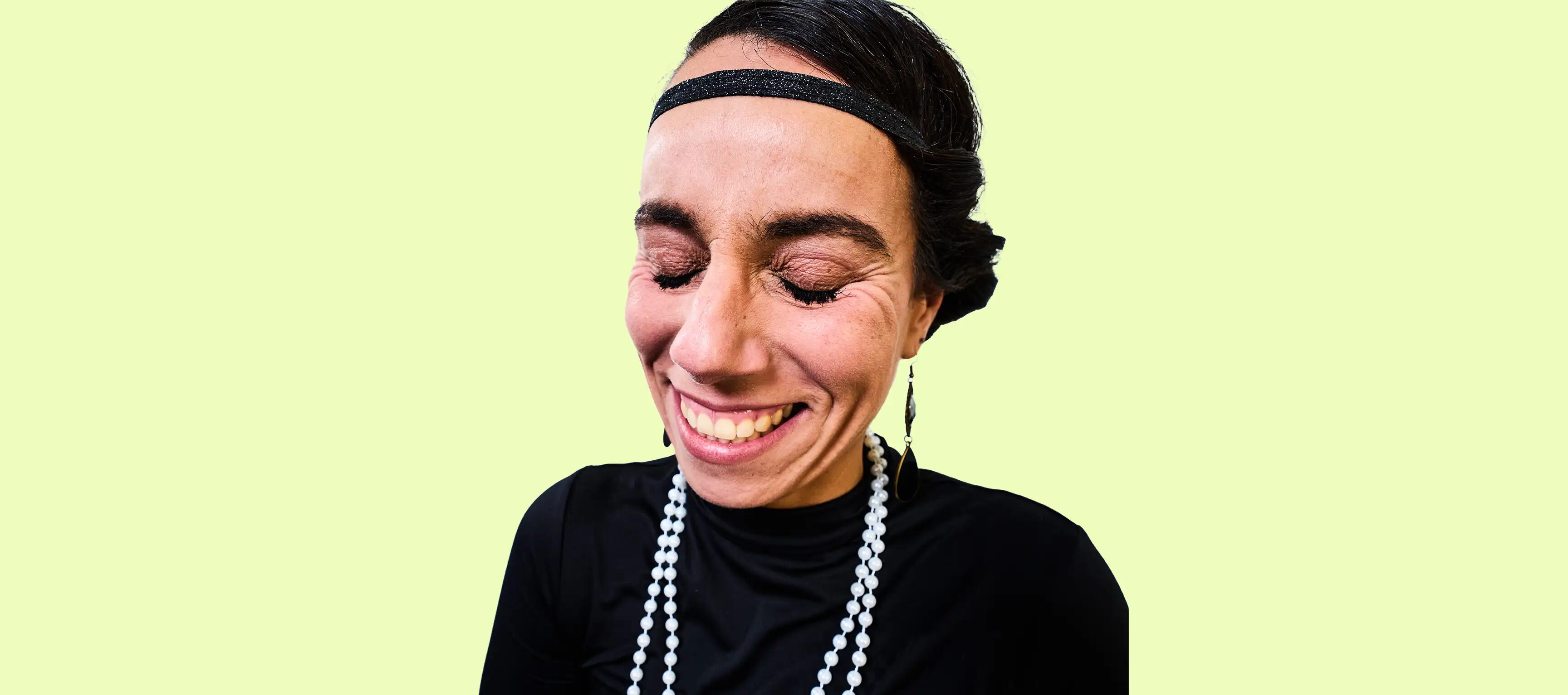 Eine lächelnde Person mit geschlossenen Augen, trägt ein schwarzes Stirnband, ein schwarzes Oberteil und eine lange, weiße Perlenkette. Der Hintergrund ist hellgrün.