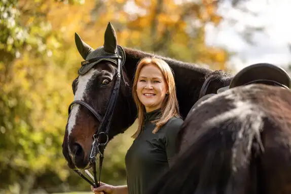 Eine lächelnde Frau steht neben einem braunen Pferd mit Zaumzeug. Sie trägt ein dunkles Oberteil. Im Hintergrund sind unscharfe Herbstbäume in Gelb- und Orangetönen zu sehen.