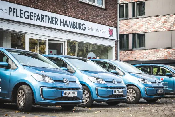 Eine Reihe von blauen Autos steht vor einem Gebäude mit der Aufschrift "Pflegepartner Hamburg" über dem Eingang. Die Fahrzeuge sind auf einem gepflasterten Parkplatz abgestellt.