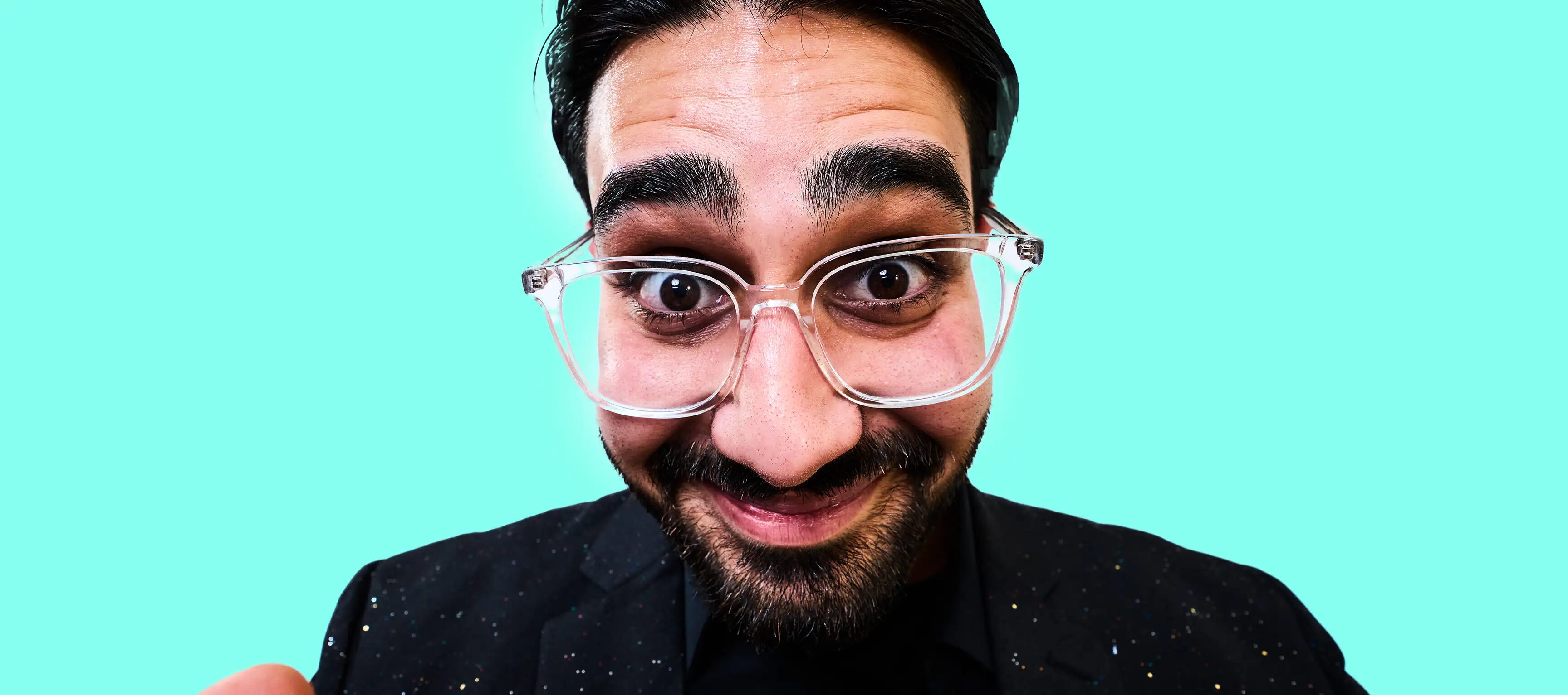 Ein lächelnder Mann mit Brille und Bart schaut direkt in die Kamera. Er trägt einen dunklen Anzug vor einem hellblauen Hintergrund.
