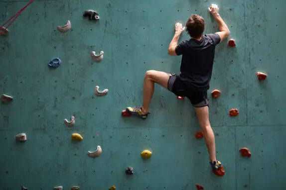 Ein Mensch klettert an einer Kletterwand mit farbigen Griffen. Die Wand ist blaugrün, und die Person trägt ein dunkles T-Shirt und Shorts. Sie ist in einer Kletterposition mit Blick auf die Wand.