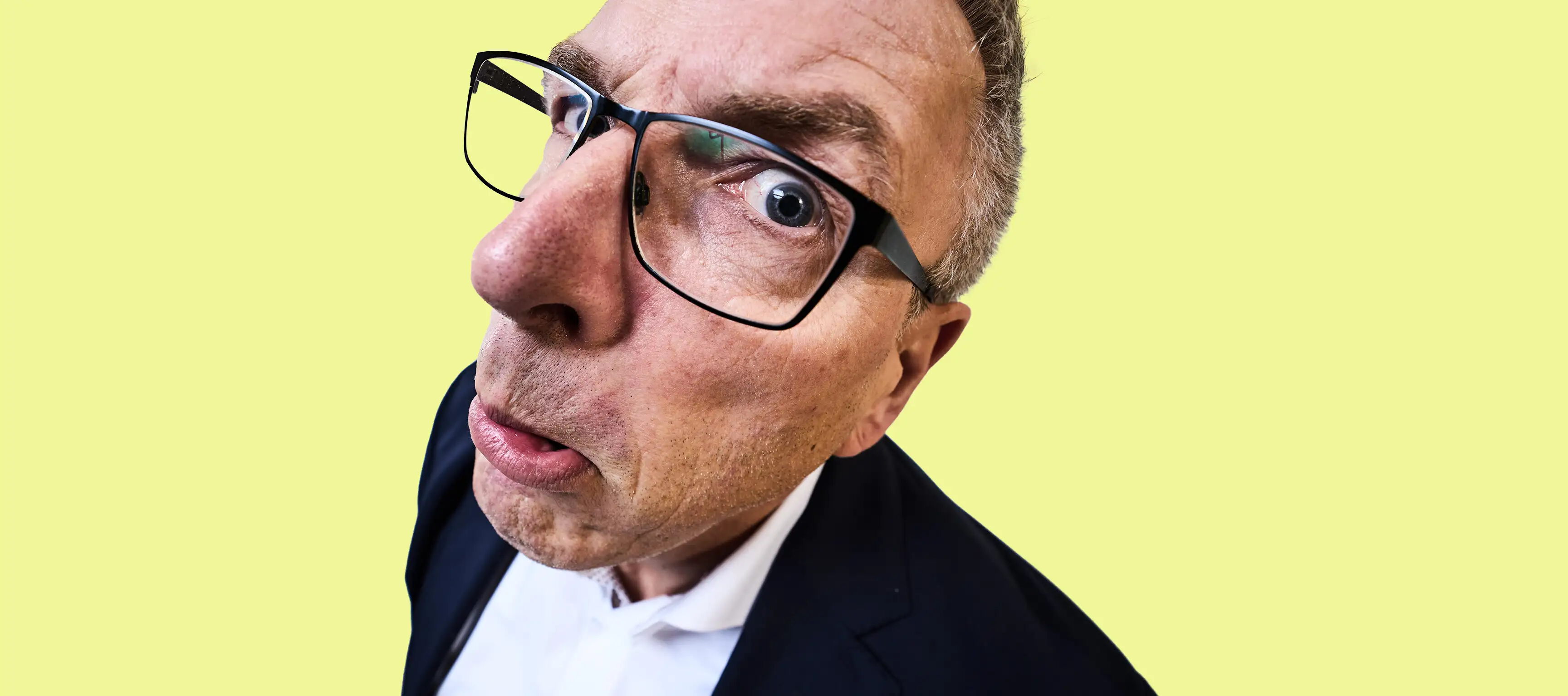 Ein Mann mit Brille und erstaunten Gesichtsausdruck schaut in die Kamera. Er trägt ein dunkles Jackett und ein weißes Hemd. Der Hintergrund ist einfarbig gelb. Die Aufnahme ist eine Nahaufnahme mit verzerrter Perspektive.