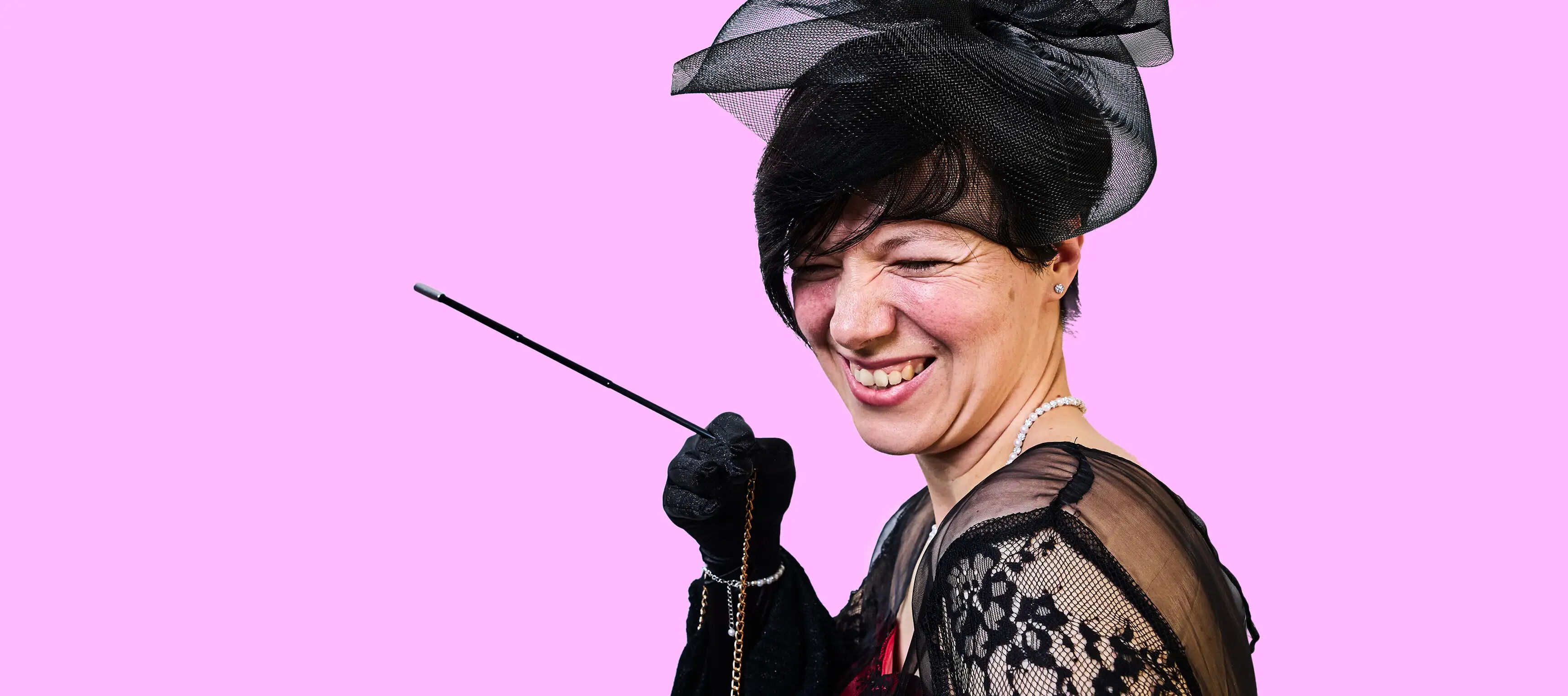 Eine Person lächelt und trägt ein schwarzes Spitzenkleid sowie einen schwarzen Hut mit Schleier. Sie hält einen schmuckartigen Stab in der Hand. Der Hintergrund ist rosa.