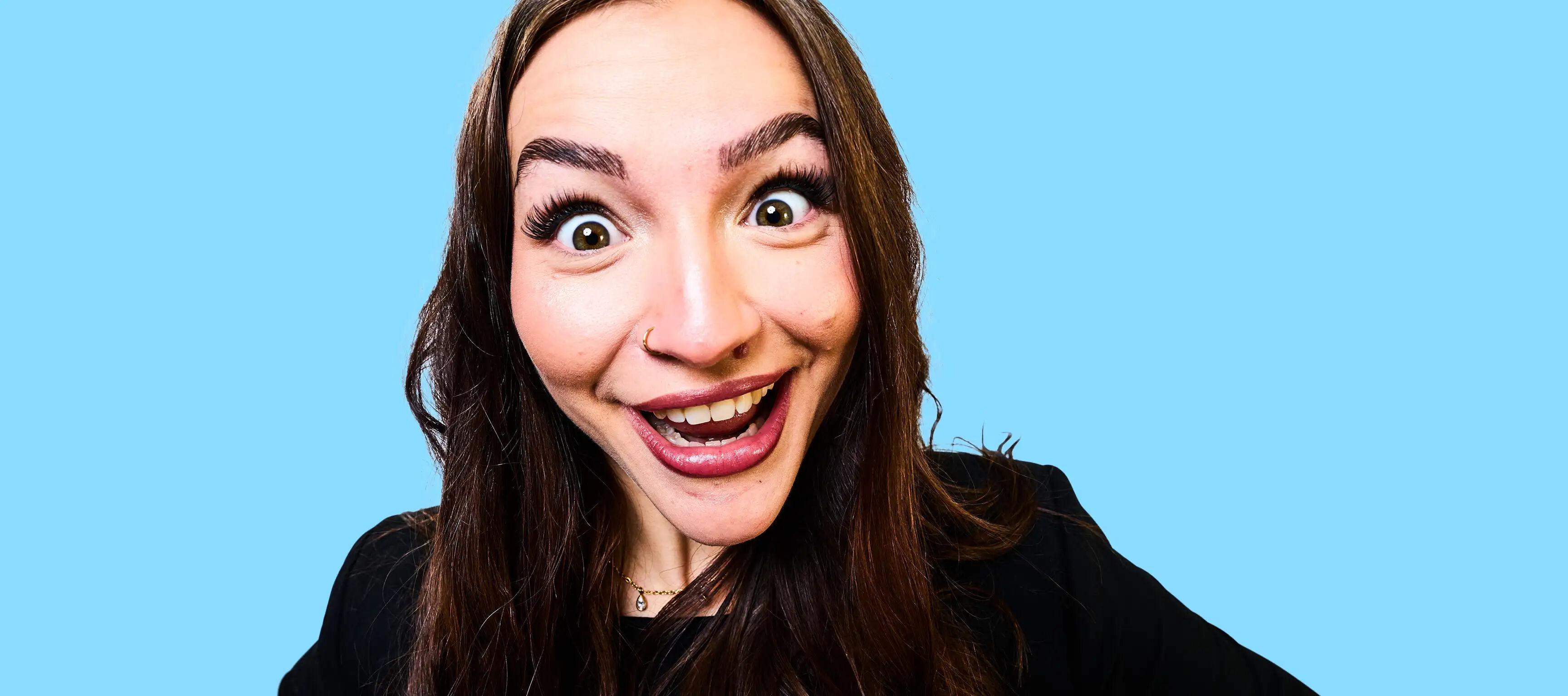 Eine Person mit langen braunen Haaren lächelt weit mit offenen Augen und leicht geöffnetem Mund, vor einem einfarbigen hellblauen Hintergrund, in die Kamera. Sie trägt dunkel Oberbekleidung.