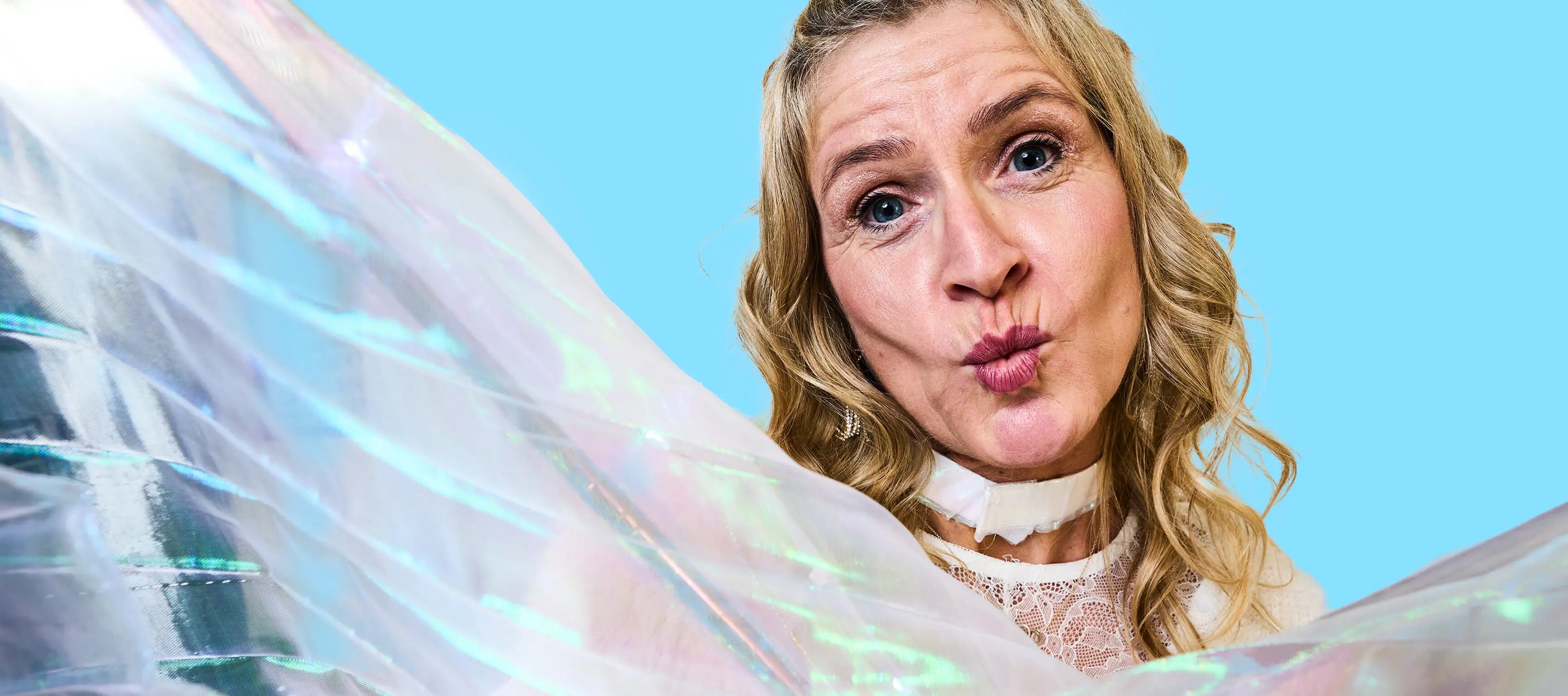 Eine Person mit langen, blonden Haaren hält ein schimmerndes, transparentes Tuch und schaut mit gespitzten Lippen in die Kamera. Der Hintergrund ist ein helles Blau.