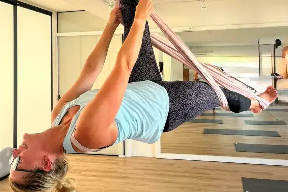 Eine Person führt eine Position in einem Aerial-Yoga-Kurs aus. Sie hängt kopfüber in einem Tuch, das an der Decke befestigt ist. Der Raum hat Holzfußboden und Spiegel an der Wand.