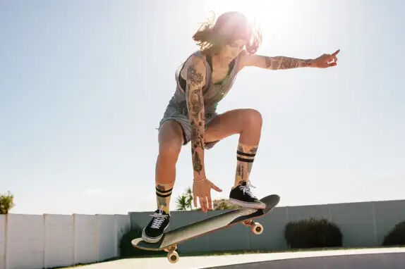 Eine Person mit Tätowierungen skatet in der Luft in einer sonnigen Umgebung, trägt einen ärmellosen Overall und schwarze Schuhe. Der Himmel ist klar und ein Zaun ist im Hintergrund zu sehen.