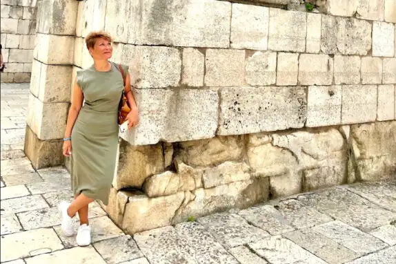 Eine Person lehnt an einer alten Steinmauer und schaut nach oben. Sie trägt ein ärmelloses, knie-langes, grünes Kleid und weiße Turnschuhe. Der Boden besteht aus unregelmäßig geformten Steinplatten.