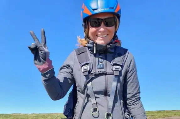 Eine Person mit blauer Helm und Sonnenbrille lächelt und zeigt das Friedenszeichen mit der Hand. Sie trägt eine graue Jacke und Handschuhe. Der Himmel ist klar und blau, und im Hintergrund ist Gras zu sehen.