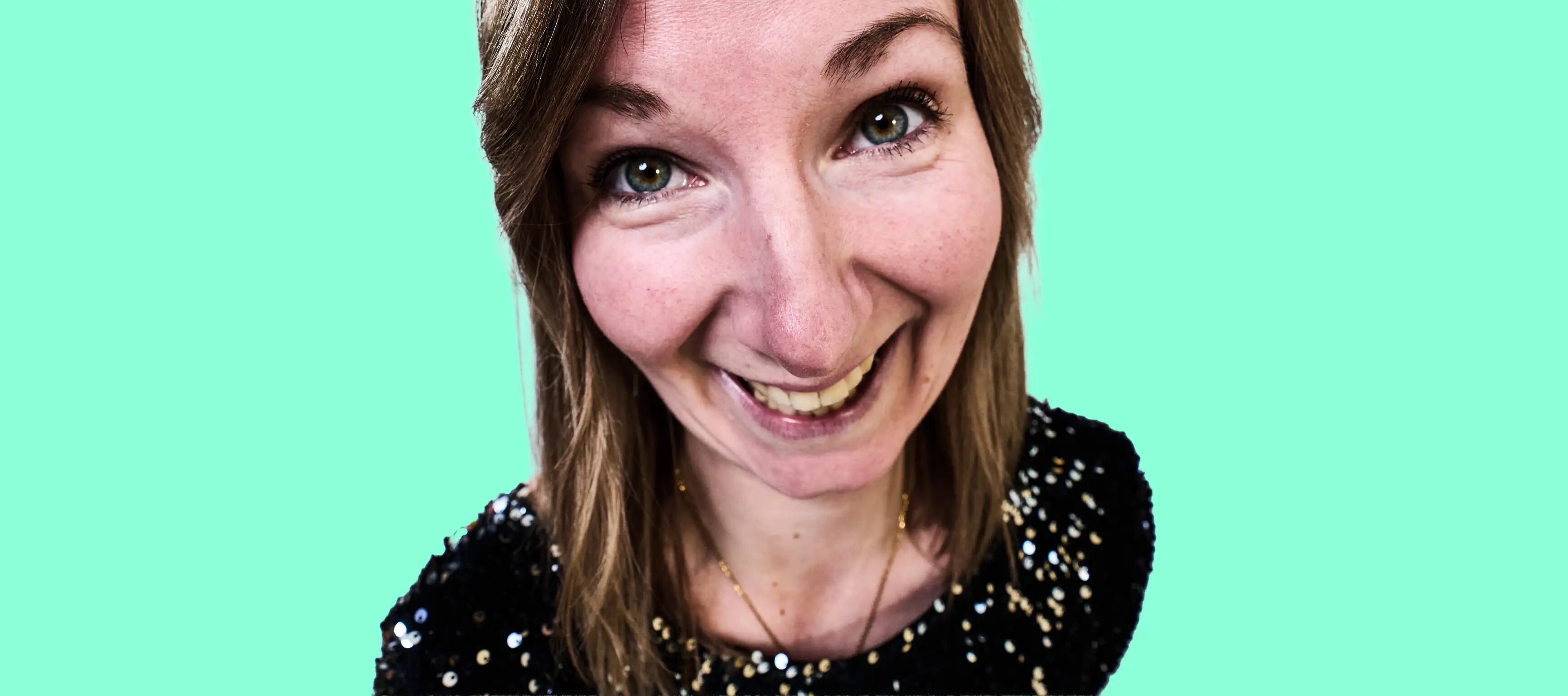 Eine Person mit mittellangem Haar und schwarzem Paillettenkleid lächelt in die Kamera. Der Hintergrund ist einfarbig hellgrün.