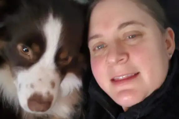 Eine Person liegt nah neben einem Hund mit braun-weißem Fell. Beide schauen in die Kamera. Der Hund hat ein flauschiges Gesicht und die Person lächelt leicht. Die Umgebung ist dunkel und unscharf.