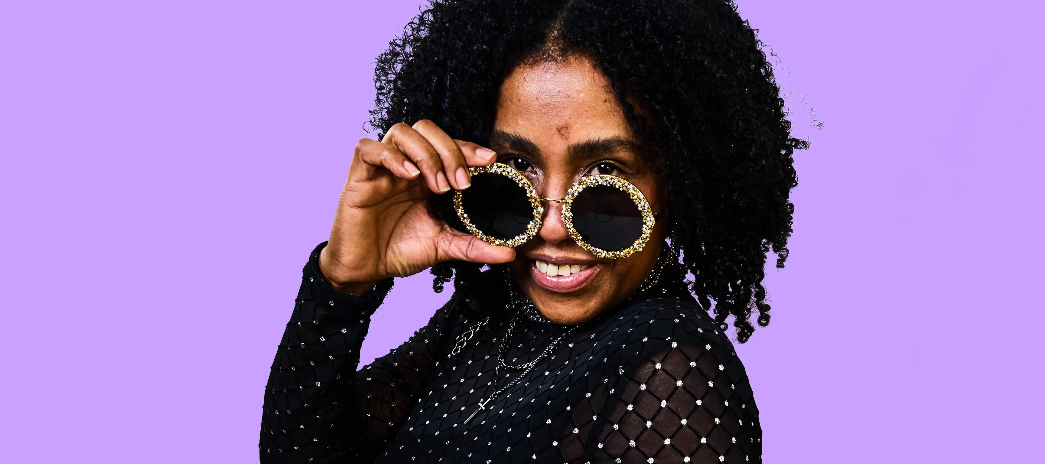 Eine Person mit lockigem Haar lächelt in die Kamera und hebt ihre goldverzierten Sonnenbrillen an. Sie trägt ein schwarzes, transparent gepunktetes Oberteil. Der Hintergrund ist einfarbig lila.