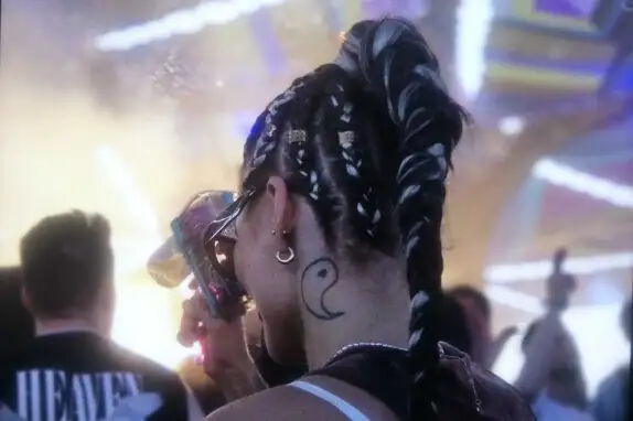 Ein Mensch mit geflochtenen Haaren, verziert mit Schmuck, hält ein buntes Getränk. Im Hintergrund sind verschwommene Lichtstrahlen und andere Personen zu sehen. Eine Tätowierung in Form eines Symbols ist am Nacken sichtbar.