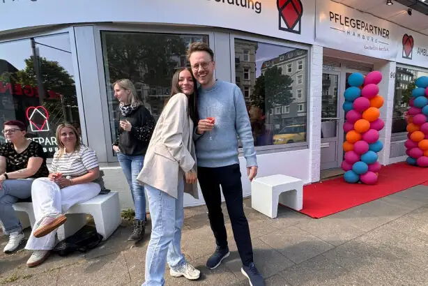 Eine Gruppe von Menschen steht vor einem Gebäude mit der Aufschrift "Pflegepartner". Zwei Personen stehen nah zusammen und lächeln. Einige sitzen auf Bänken. Bunte Luftballons schmücken den Eingang. Es ist tagsüber.