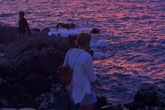 Zwei Personen stehen auf Felsen am Meer und blicken auf das Wasser. Der Himmel ist in rosa und violetten Tönen gefärbt, was sich im Wasser spiegelt. Eine Person trägt eine weiße Jacke und einen Rucksack.