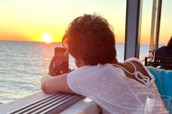 Eine Person lehnt sich an ein Geländer und fotografiert mit einem Smartphone einen Sonnenuntergang über dem Meer. Die Sonne taucht den Himmel in warme Farben. Im Vordergrund sind ein Tisch und ein Getränkeglas zu sehen.