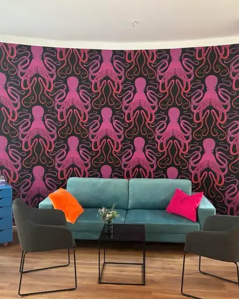 Ein Wohnzimmer mit einer auffälligen Tapete, die pinke Oktopusmuster auf schwarzem Hintergrund zeigt. Im Raum stehen ein blaues Sofa mit bunten Kissen, zwei schwarze Sessel und ein kleiner Tisch.