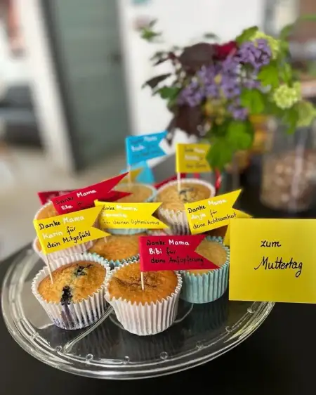 Ein Tablett mit sieben Muffins, die jeweils bunte Fähnchen mit Danksagungen tragen. Im Hintergrund steht eine Vase mit Blumen. Daneben ein gelber Zettel mit der Aufschrift "zum Muttertag".