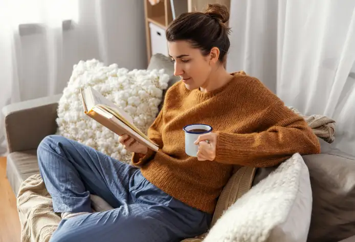 Eine Person sitzt entspannt auf einem Sofa, trägt einen braunen Pullover und blaue Hose und liest ein Buch. In der anderen Hand hält sie eine Tasse. Um sie herum sind Kissen und ein heller Vorhang im Hintergrund.