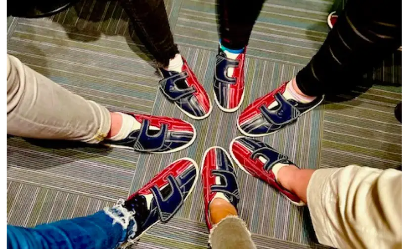 Fünf Personen stehen im Kreis und zeigen ihre identischen Schuhe. Die Schuhe sind dunkelblau und rot mit einem markanten weißen Muster. Der Boden hat ein gestreiftes Muster in verschiedenen Grautönen.