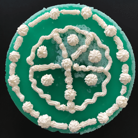Ein runder, grüner Kuchen mit weißer Sahneverzierung. Die Verzierung besteht aus einem äußeren Kreis, einem inneren Muster mit vier Abschnitten und einem kleineren Kreis in der Mitte, jeweils verziert mit Sahnetuffs.