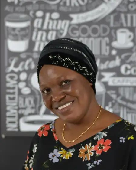 Eine lächelnde Person trägt ein schwarzes Kopftuch mit silbernen Mustern und ein schwarzes Oberteil mit bunten Blumen. Der Hintergrund zeigt weiße, stilisierte Schriftzüge und Kaffeetassenmotive auf einer dunklen Fläche.