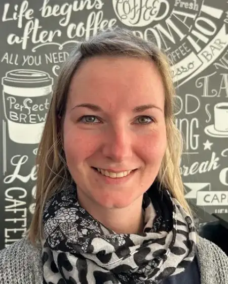 Das Bild zeigt eine lächelnde Person vor einer Wand mit Kaffeemotiven und Schriftzügen. Die Person trägt einen gemusterten Schal.