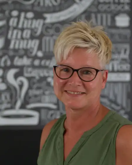 Eine Person mit kurzen, blonden Haaren und Brille lächelt. Sie trägt ein ärmelloses, grünes Oberteil. Der Hintergrund zeigt eine Wand mit Kreideschrift und stilisierten Zeichnungen in Schwarz-Weiß.