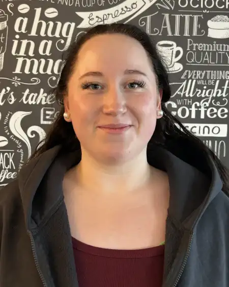 Eine Person mit langen Haaren trägt eine Kapuzenjacke und lächelt leicht in die Kamera. Der Hintergrund ist eine Wand mit weißem Text und Kaffeetassenmotiven auf schwarzem Grund.